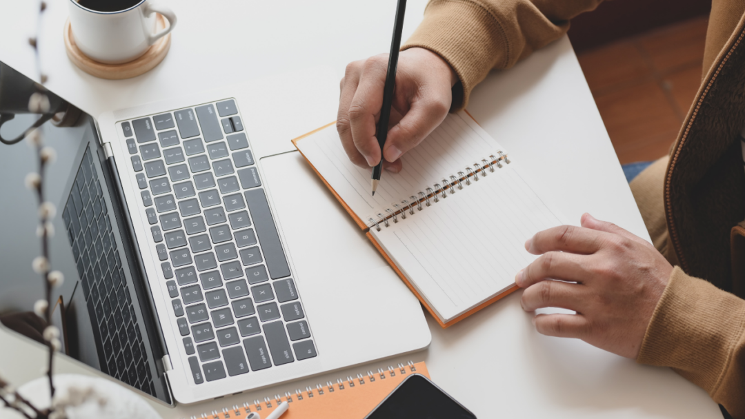 Worker at desk writing in notebook by open laptop